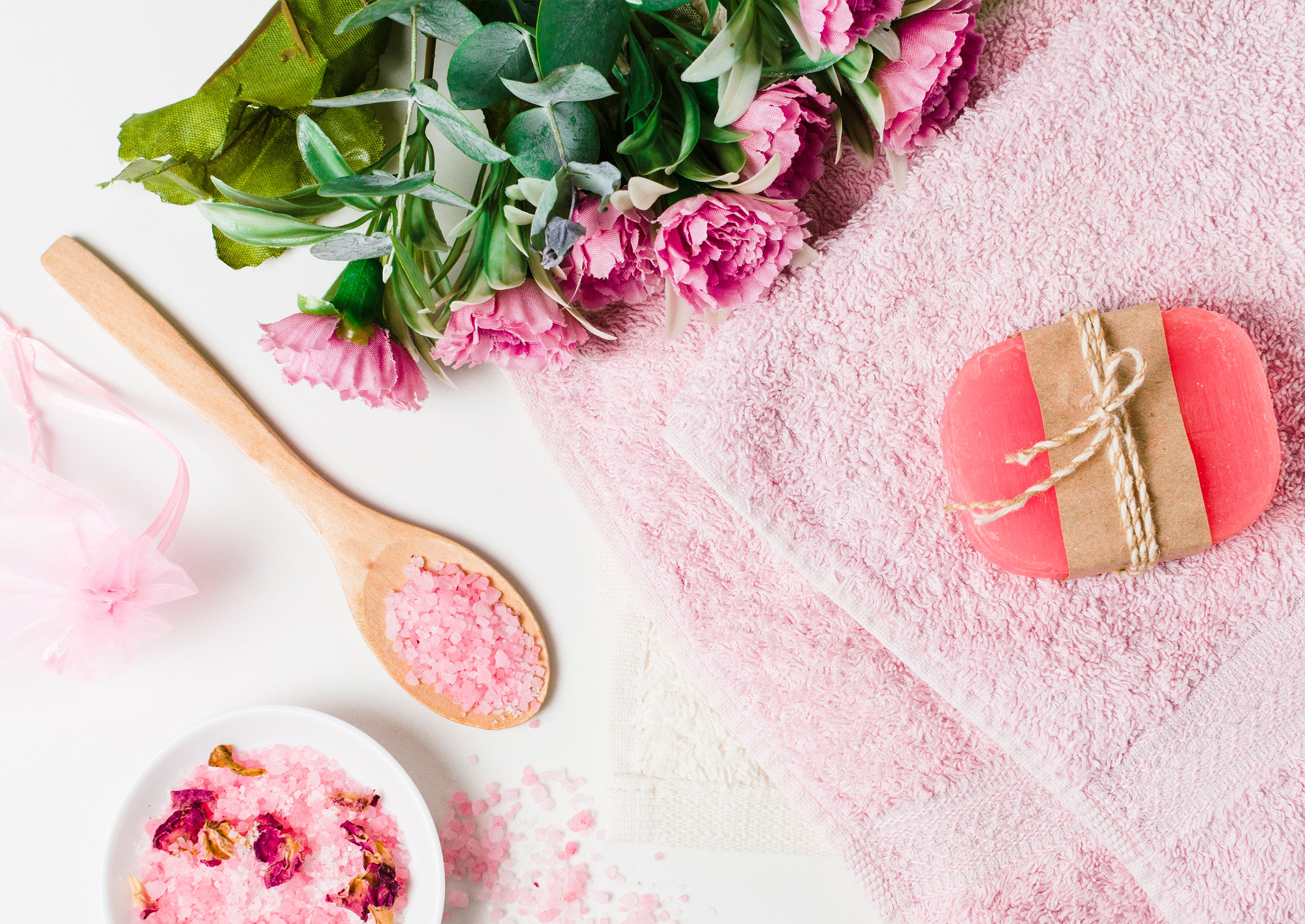 Il s'agit d'une image avec des affaires de bains : un savon rose, une serviette blanche, du sel de bain rose dans récipients en porcelaine et une cuillère en bois, un bouquet d'oeillets rose