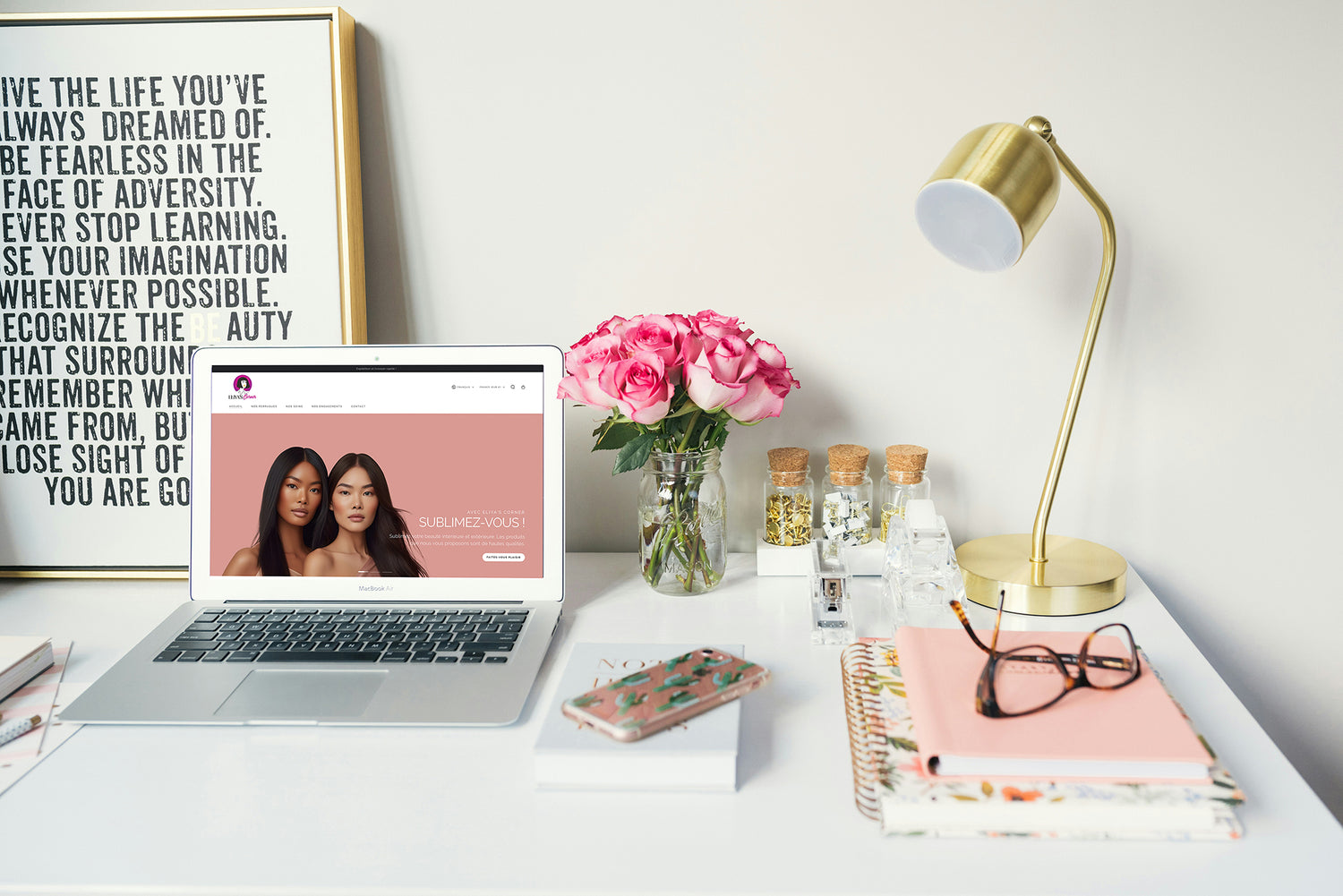 Image d'un ordinateur portable avec la boutique Eliyascorner sur l'Écran. Un bureau très féminin avec des livres, un bouquets de rose dans un pot en verre, une lampe dorée, un téléphone portable.